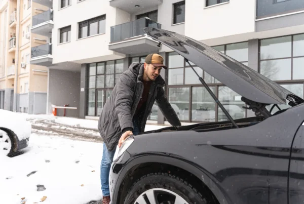 A man in a winter coat and cap leans over the open hood of a black SUV parked in the snow, looking into the engine bay.