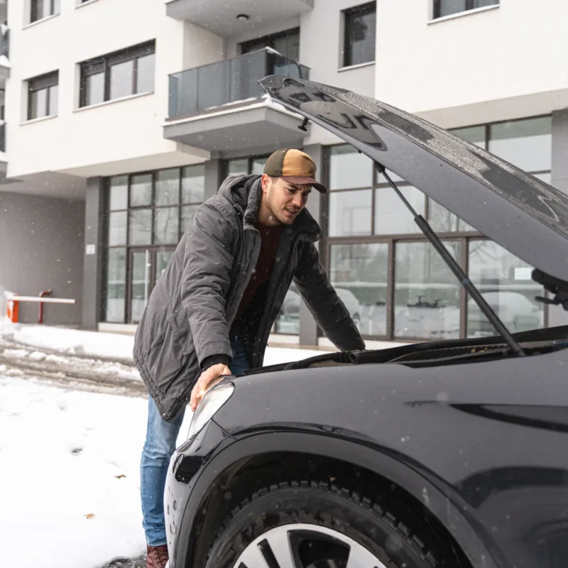 A man in a winter coat and cap leans over the open hood of a black SUV parked in the snow, looking into the engine bay.