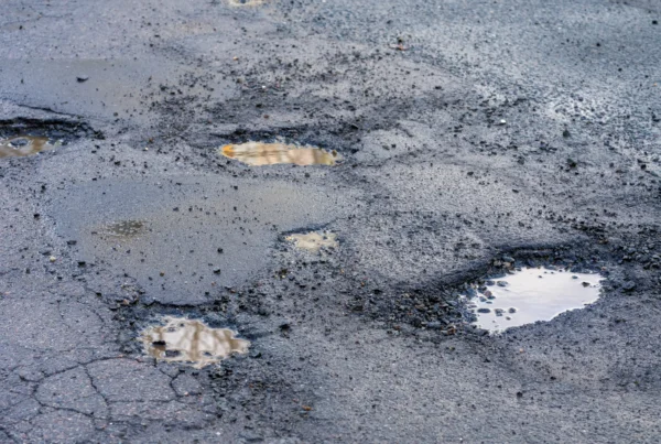 A cracked asphalt road surface with several potholes filled with muddy water and loose gravel around their edges.