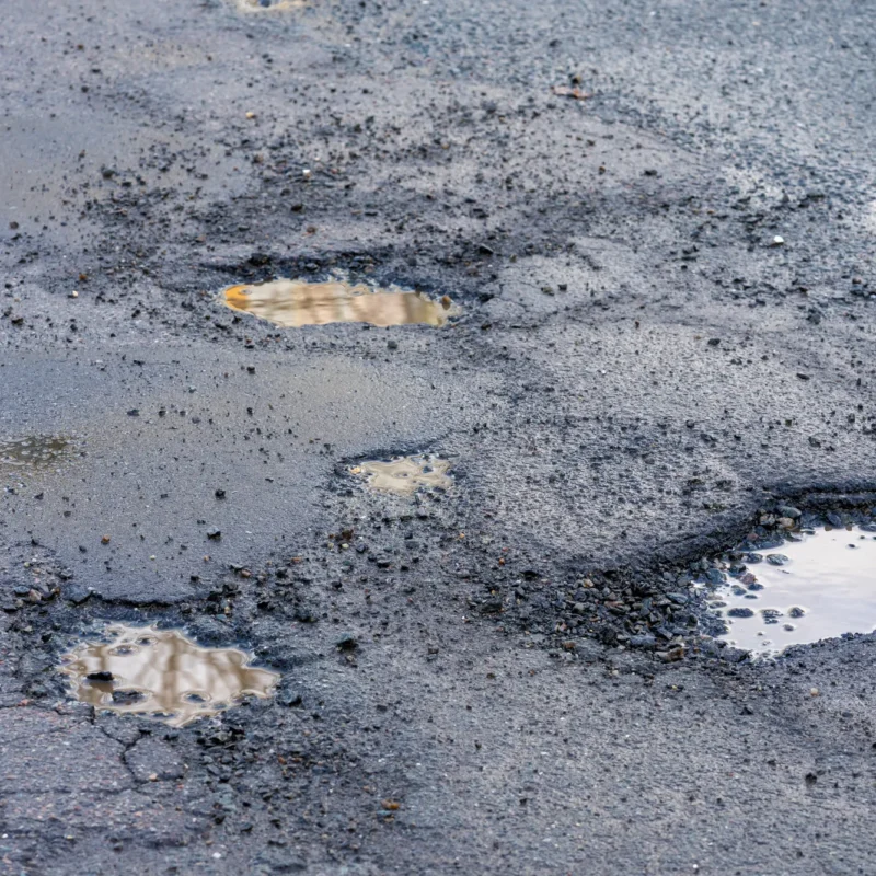 A cracked asphalt road surface with several potholes filled with muddy water and loose gravel around their edges.