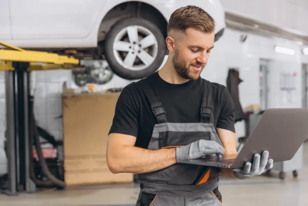 A smiling auto technician in gray overalls uses a laptop in a repair shop while a white car sits raised on a lift behind him.