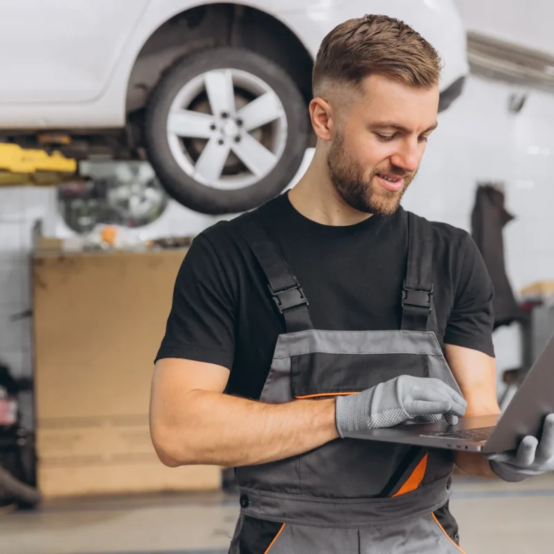 A smiling auto technician in gray overalls uses a laptop in a repair shop while a white car sits raised on a lift behind him.