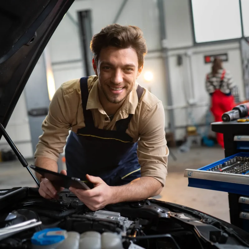 A smiling mechanic in overalls holds a tablet while leaning over the open hood of a car inside an auto repair shop.