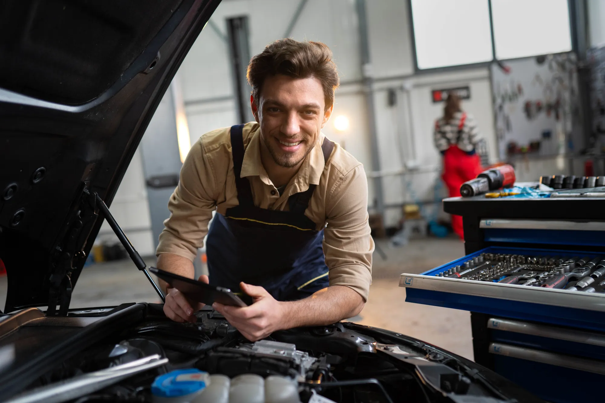 A smiling mechanic in overalls holds a tablet while leaning over the open hood of a car inside an auto repair shop.
