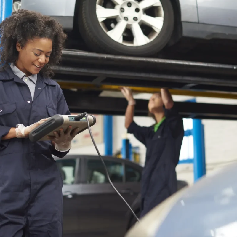 A female mechanic in navy coveralls reads a handheld diagnostic tool while a coworker works beneath a car raised on a lift in a repair shop.