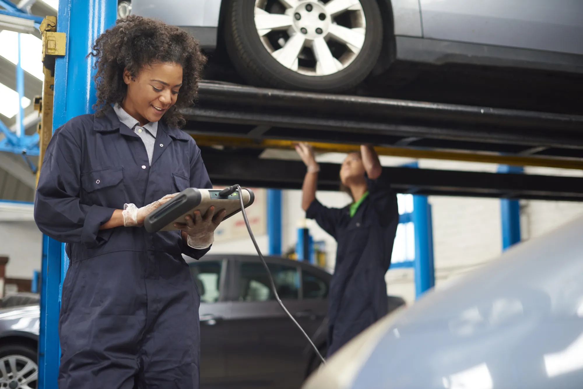 A female mechanic in navy coveralls reads a handheld diagnostic tool while a coworker works beneath a car raised on a lift in a repair shop.