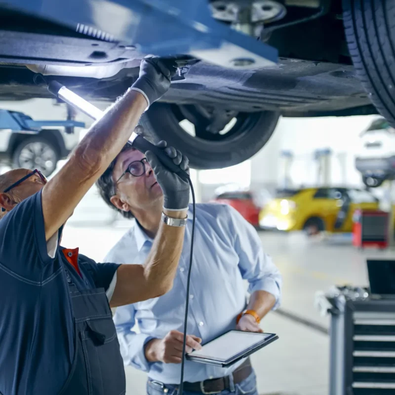 A mechanic shines a work light on the underside of a car raised on a lift while a man holding a tablet looks up at it.