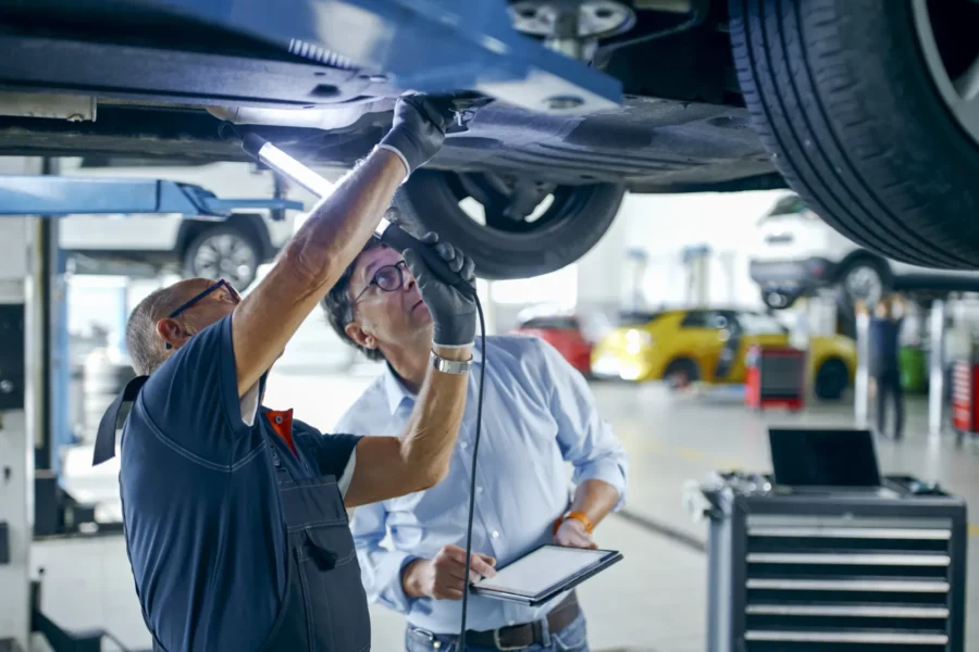 A mechanic shines a work light on the underside of a car raised on a lift while a man holding a tablet looks up at it.