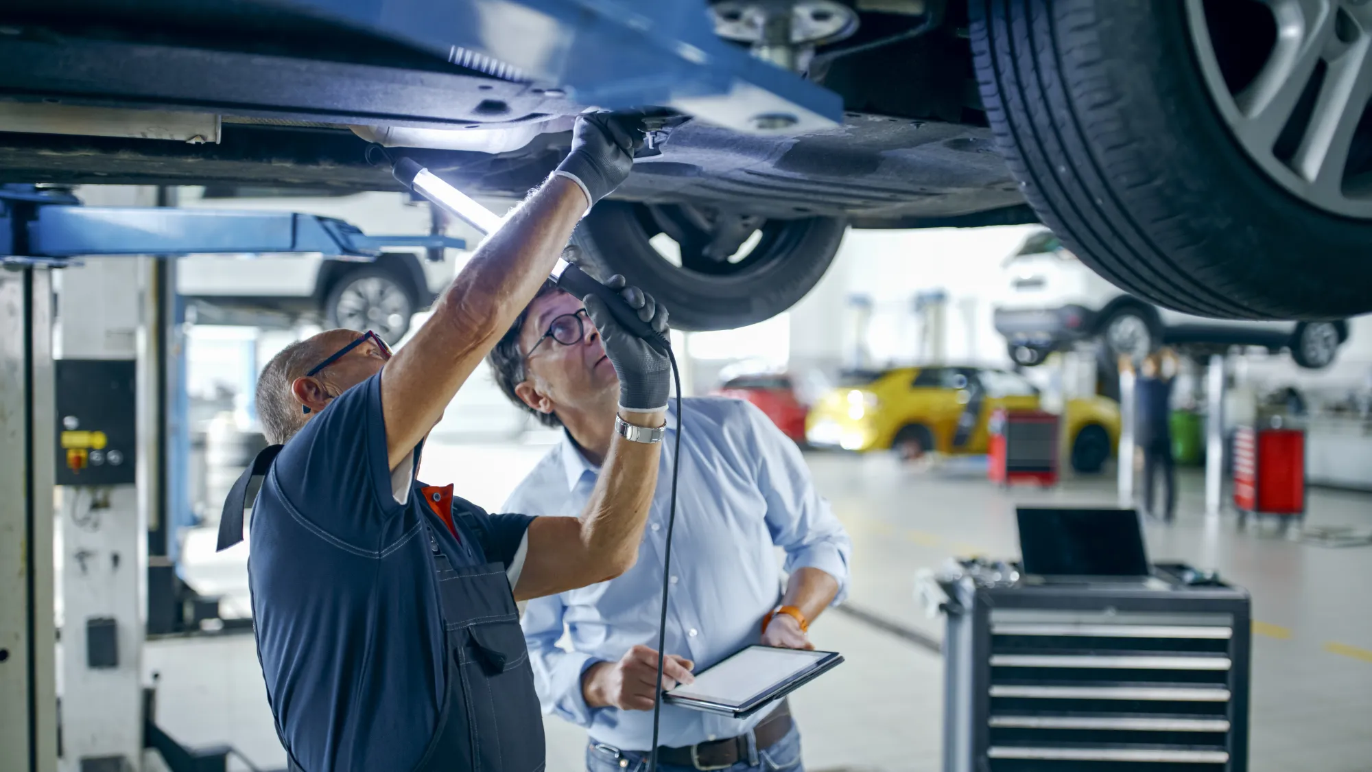 A mechanic shines a work light on the underside of a car raised on a lift while a man holding a tablet looks up at it.
